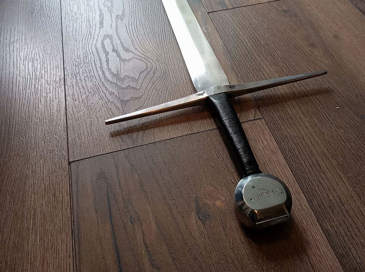 Gevest van een handgesmeed langzwaard op een donkere houten vloer: pareerstang, leren greep en pommel met smidsmerk